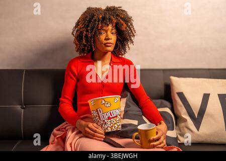 Young woman sitting on sofa with popcorn bucket and coffee mug, enjoying a movie night at home Stock Photo
