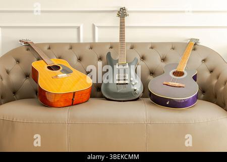 Three guitars set on a brown leather sofa in the living room. A black electric guitar, a brown acoustic and a purple classical guitar. Indoors at home Stock Photo