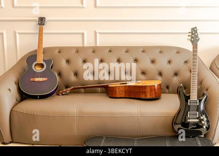 Three guitars set on a brown leather sofa in the living room. A black electric guitar, a brown acoustic and a purple classical guitar. Indoors at home Stock Photo