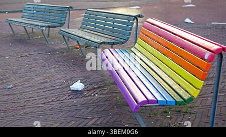 Colorful rainbow bench in urban park. A vibrant rainbow-colored bench stands out among standard benches in a brick-paved urban park, surrounded by Stock Photo