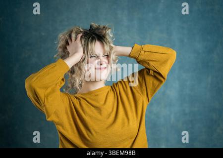 A young woman in a yellow sweater expresses frustration by clutching her curly hair against a textured blue backdrop. Her expression captures a moment Stock Photo