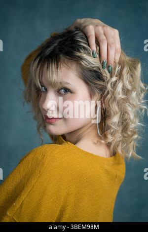 Portrait of a young woman with curly hair, wearing a yellow sweater and hoop earrings. She poses confidently with her hand in her hair. Blue backgroun Stock Photo