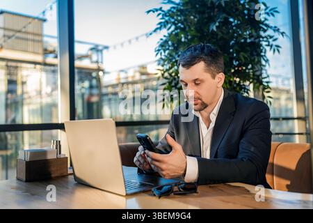 A businessman in a suit works with a laptop at a cafe table. The environment is relaxed, with natural light streaming through large windows, creating Stock Photo