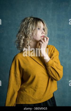A contemplative young woman with curly blond hair, wearing a yellow sweater, poses thoughtfully against a textured blue background, highlighting a mom Stock Photo