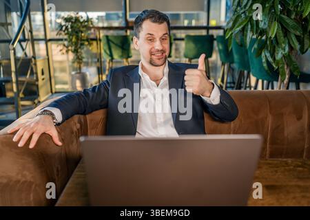 A smiling businessman sits with a laptop in a cozy cafe, giving a thumbs-up gesture. Dressed in a dark suit and seated on a sofa, he appears upbeat, s Stock Photo