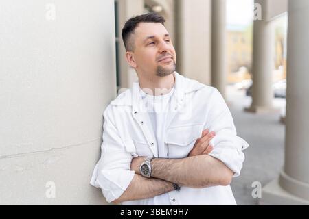 A contemplative male businessman in casual white attire, leaning against a building pillar in an urban setting, appears pensively lost in thought. Stock Photo