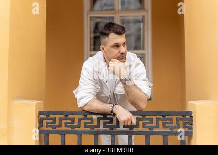A young businessman in elegant white attire contemplates while leaning on an ornate balcony railing, set against a soft yellow building backdrop. Stock Photo