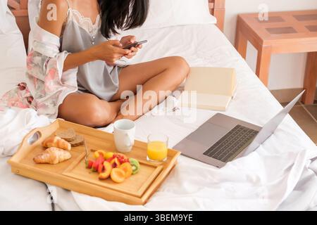 Crop unrecognizable female in nightwear messaging on cellphone while sitting near laptop and open book on bed and having breakfast in morning Stock Photo