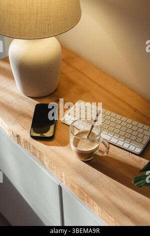 A cozy home workspace featuring a warm lamp, a clear glass of coffee, a smartphone, and a keyboard on a wooden desk, creating a serene and productive Stock Photo