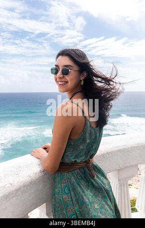 A cheerful young woman in a green dress and sunglasses smiles while looking at the Caribbean Sea from a coastal balcony in Cozumel, Mexico. Stock Photo