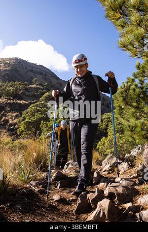 Enthusiastic hikers navigate rugged trails in sunny weather, wearing helmets and using poles. Framed by green foliage, they embody adventure and the g Stock Photo