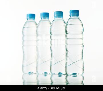 Four clear plastic water bottles with blue caps stand in a straight line, casting soft reflections on the glossy surface beneath. The backdrop is a cl Stock Photo