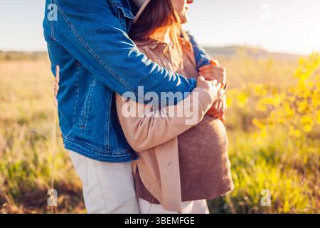 Pregnancy. Pregnant family couple walking on spring meadow at sunset. Husband hugging his wife from behind. Close up Stock Photo