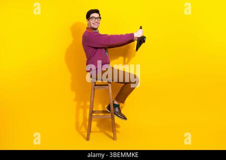Young man with glasses performing a humorous driving gesture on a stool against a vibrant yellow background Stock Photo