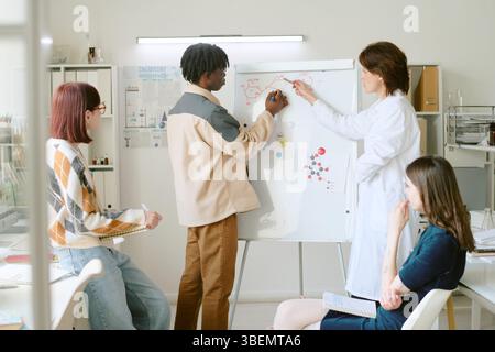 Group of multiracial classmates collaborating on project, drawing on whiteboard and discussing research in contemporary lab setting Stock Photo