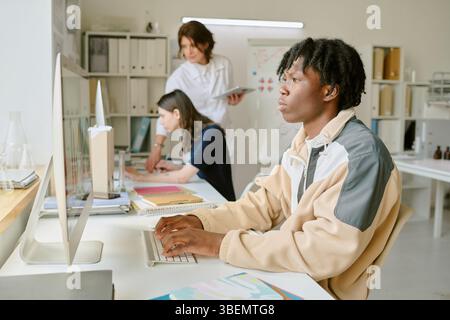 Student working on computer, focused and typing while two colleagues collaborate in background in bright, organized office space featuring various wor Stock Photo