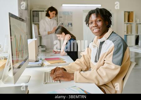 Smiling African American student working on computer in modern lab with classmates in background engaging in various tasks. Visible computer screen sh Stock Photo