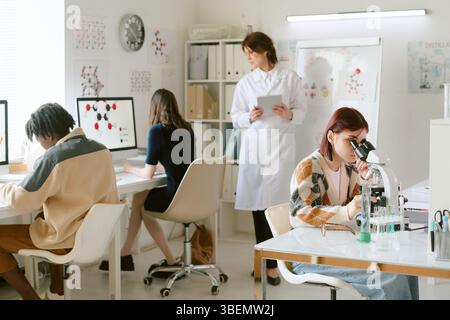Group of researchers engaged in various scientific activities at laboratory with one woman using microscope in forefront and others analyzing data on Stock Photo