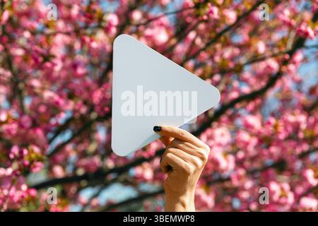 Hand holding a white play button icon in front of blooming cherry blossom tree on a bright spring day. Stock Photo