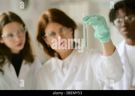 Female scientist with multiracial team wearing lab coats and protective eyewear analyzing chemical reactions in science laboratory while holding test Stock Photo