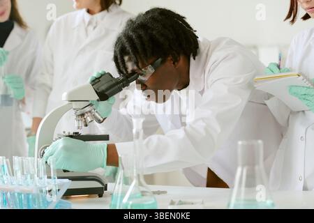 African American student examining samples under microscope while classmates taking notes in laboratory environment with lab equipment and glassware Stock Photo