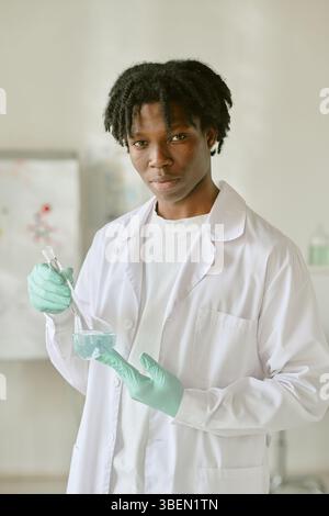 Portrait of African American student wearing lab coat and gloves, mixing chemicals in laboratory setting. Scientific equipment and charts in backgroun Stock Photo