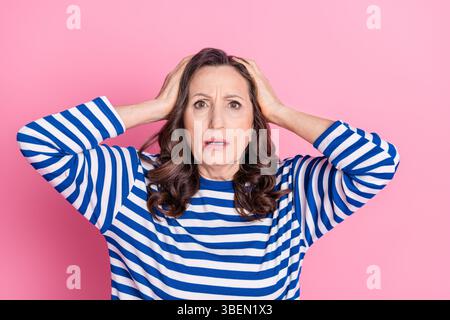 Surprised mature woman in striped sweater against pink background expressing emotion with hands on head Stock Photo