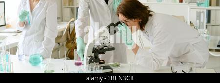 Group of scientists conducting experiments in laboratory, examining samples under microscope while wearing white lab coats and green gloves Stock Photo
