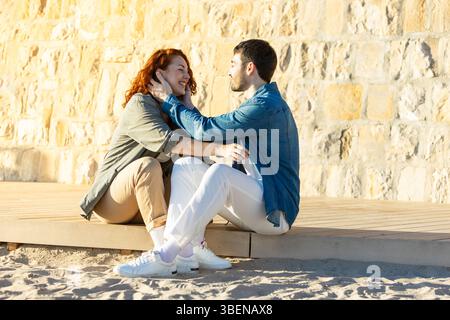Young couple sitting closely on a wooden boardwalk, smiling as the man gently holds the woman s face in his hands, surrounded by golden light and a st Stock Photo
