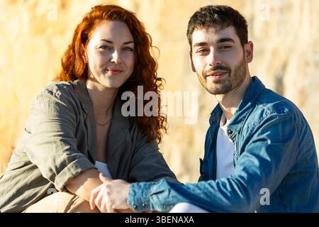 Close-up of a young couple holding hands and smiling at the camera while sitting outdoors, with golden sunlight illuminating their faces against a sto Stock Photo