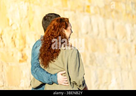 Rear view of a couple hugging closely and looking forward together, bathed in golden sunlight with a soft-focus stone wall in the background. Lifestyl Stock Photo
