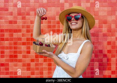 Influencer showing a basket of cherries while holding two cherries with two fingers in front of a red tiled wall Stock Photo
