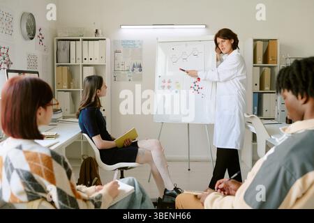 Female scientist explaining chemistry concepts to diverse group of students in modern classroom setting while using whiteboard and diagrams Stock Photo