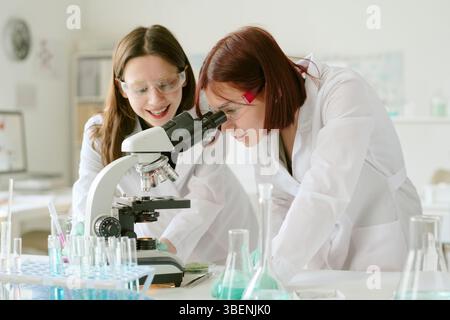 Two young female scientists working together in laboratory, using microscope to analyze samples while wearing protective eyewear, surrounded by lab eq Stock Photo