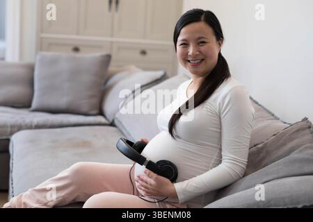 Asian pregnant woman sitting on sofa holding black over-ear headphones on belly at home, copy space Stock Photo