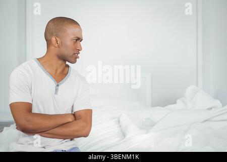 African American man sitting on edge of unmade bed in bedroom with white pillows, copy space Stock Photo