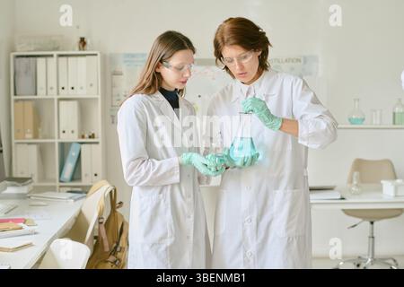 Two scientists in lab coats and gloves examining blue liquid in laboratory. Laboratory equipment and glassware organized neatly in background Stock Photo