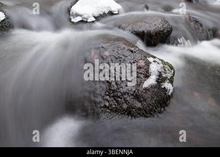 Smooth dark rocks sit in a gently flowing winter river, partially covered in thin layers of snow and ice. Stock Photo