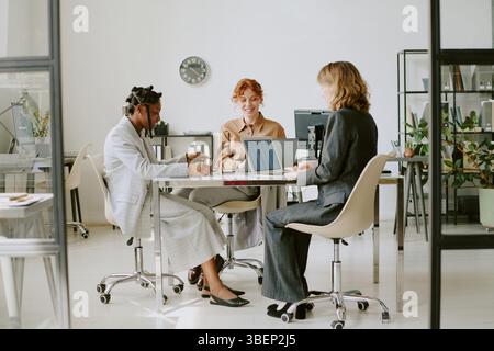 Three professionals sitting around office table, engaging in serious discussion, with laptops and documents. Modern office setup with glass walls and Stock Photo