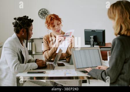 Multiracial team collaborating on project with laptops and documents on modern office desk, engaging in discussion for strategy and planning Stock Photo