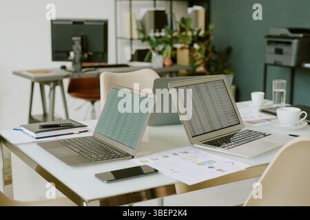 Office desk filled with laptops, documents and coffee cups, showing productive work environment with charts and computer screens displaying data in ba Stock Photo