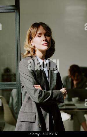 Portrait of confident businesswoman with blond hair and folded arms standing in modern office with colleague working in background. Direct sunlight cr Stock Photo