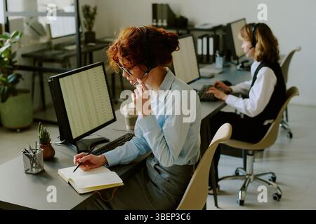 Two office workers focused on tasks at their desks, with one taking notes and wearing headset. Modern workspace with computers and plants creating pro Stock Photo