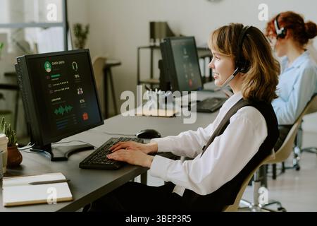 Woman with headset working on computer in modern office environment with other colleagues in background, focusing on tasks and using technology effici Stock Photo
