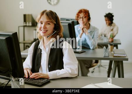 Portrait of customer support representative wearing headset sitting at desk smiling, with colleagues working at computers in office background Stock Photo