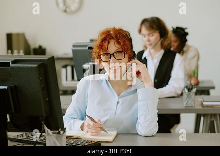 Portrait of red-haired woman wearing glasses and writing notes while talking on phone in a modern office. Co-workers visible in background working dil Stock Photo