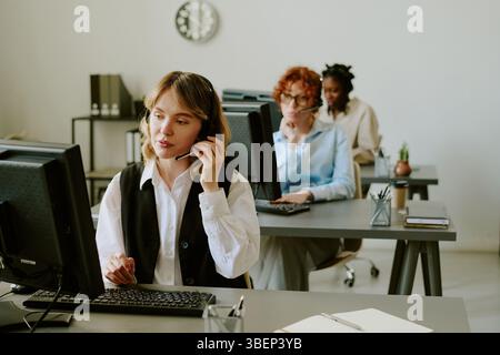 People focused on work in office with computers and phones on their desks. One person using phone, while others working on their computers Stock Photo