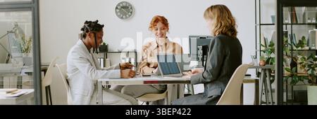 Three professionals engaged in collaborative meeting with laptops and documents on table. Diverse group working on project in spacious, well-lit offic Stock Photo