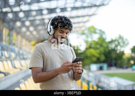A smiling man in sportswear wearing headphones, holding a smartphone while standing in a stadium setting, outdoors. The background is blurred. Stock Photo