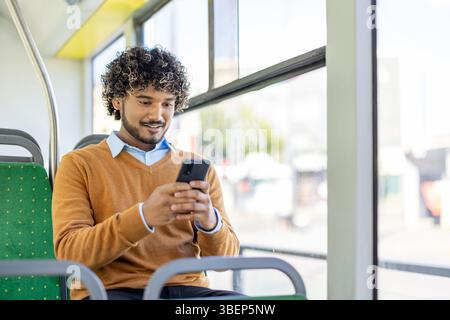 A smiling man using a smartphone while seated inside a public transportation vehicle, near a window. Stock Photo